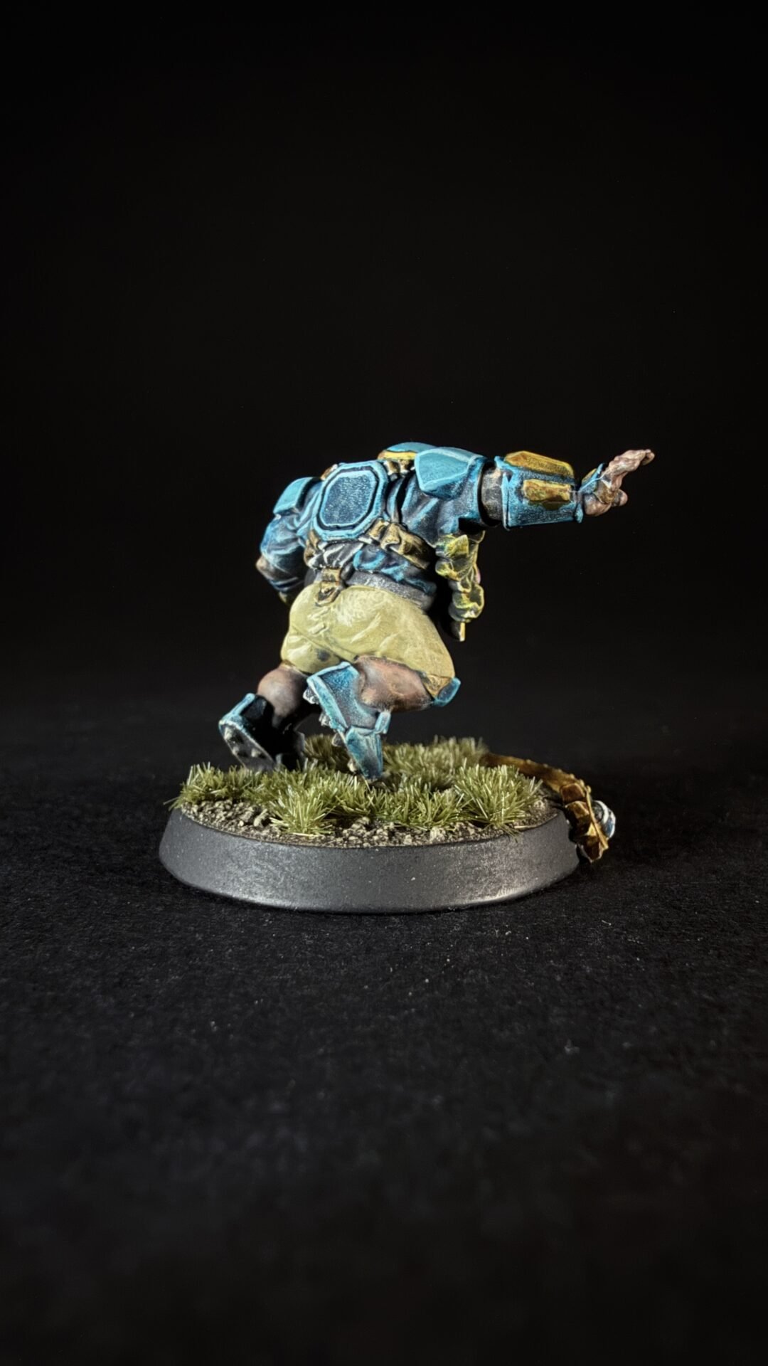 Miniature sci-fi soldier in blue armor leaping forward on a grassy base, viewed from the side against a dark backdrop.
