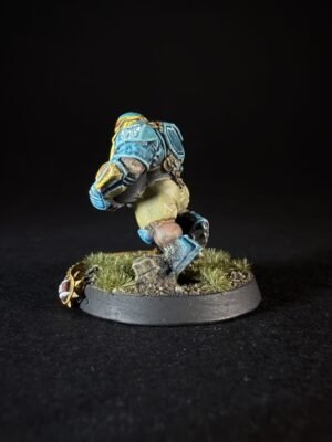 Miniature sci-fi soldier in blue armor running on a grassy diorama base, with dirt and tufts of grass under a black backdrop.