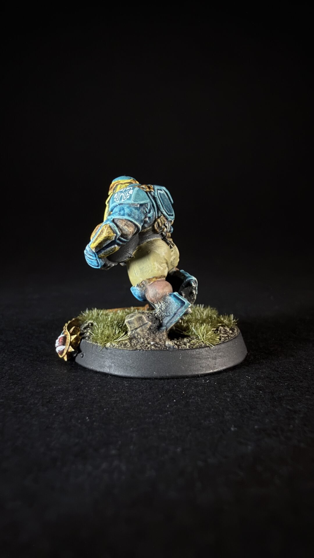 Miniature sci-fi soldier in blue armor running on a grassy diorama base, with dirt and tufts of grass under a black backdrop.
