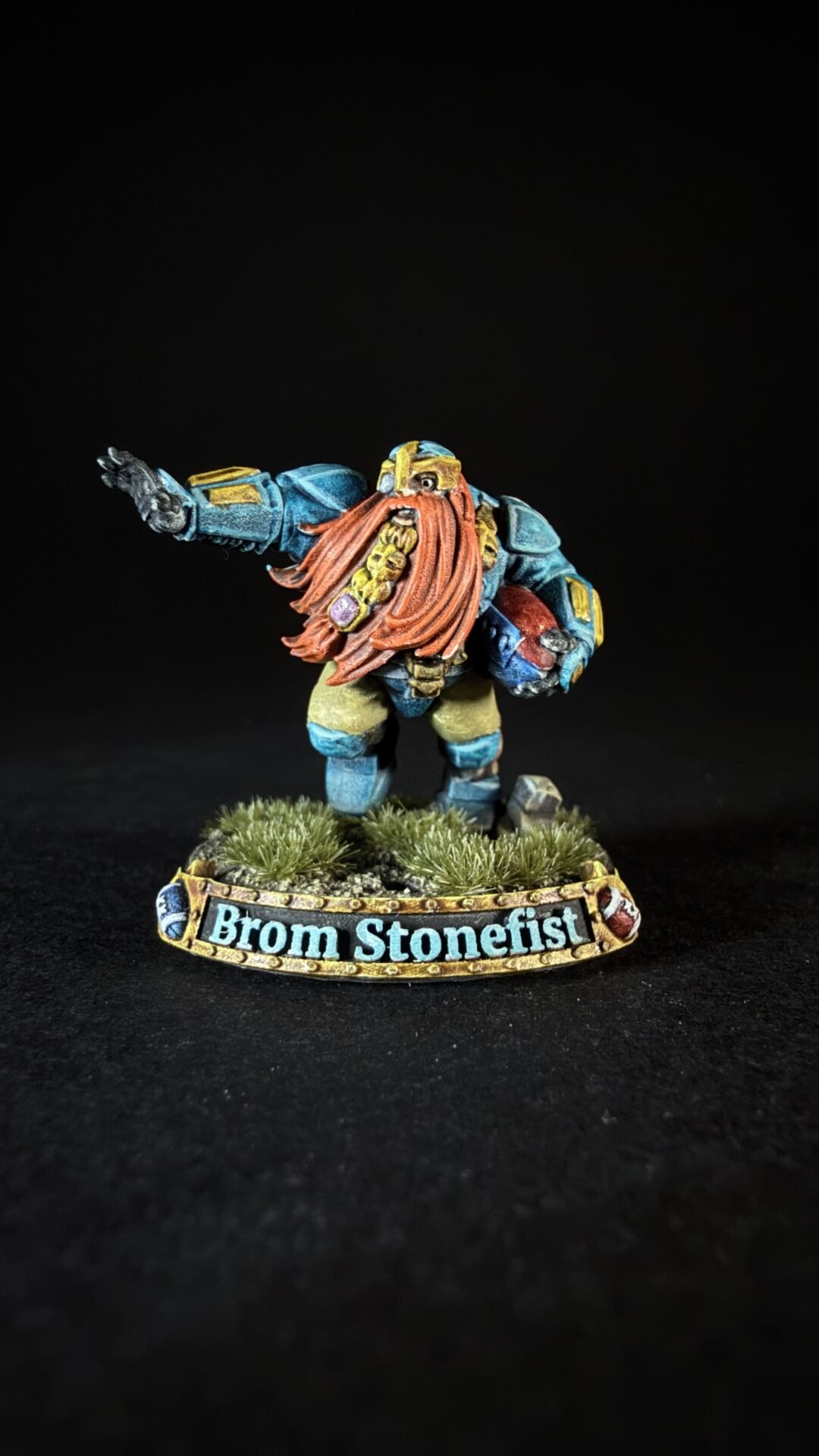 Miniature dwarf warrior in blue armor with a long orange beard, standing on a grassy base with a 'Brom Stonefist' name banner on the base