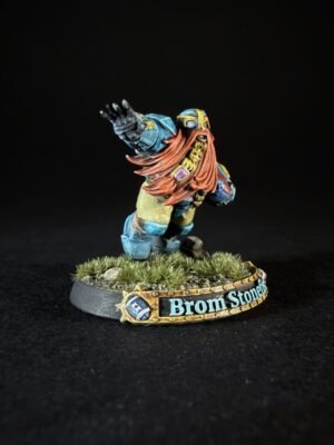 Miniature dwarf warrior with long red beard and blue armor raises his arm on a grassy base labeled Brom Stonehide.