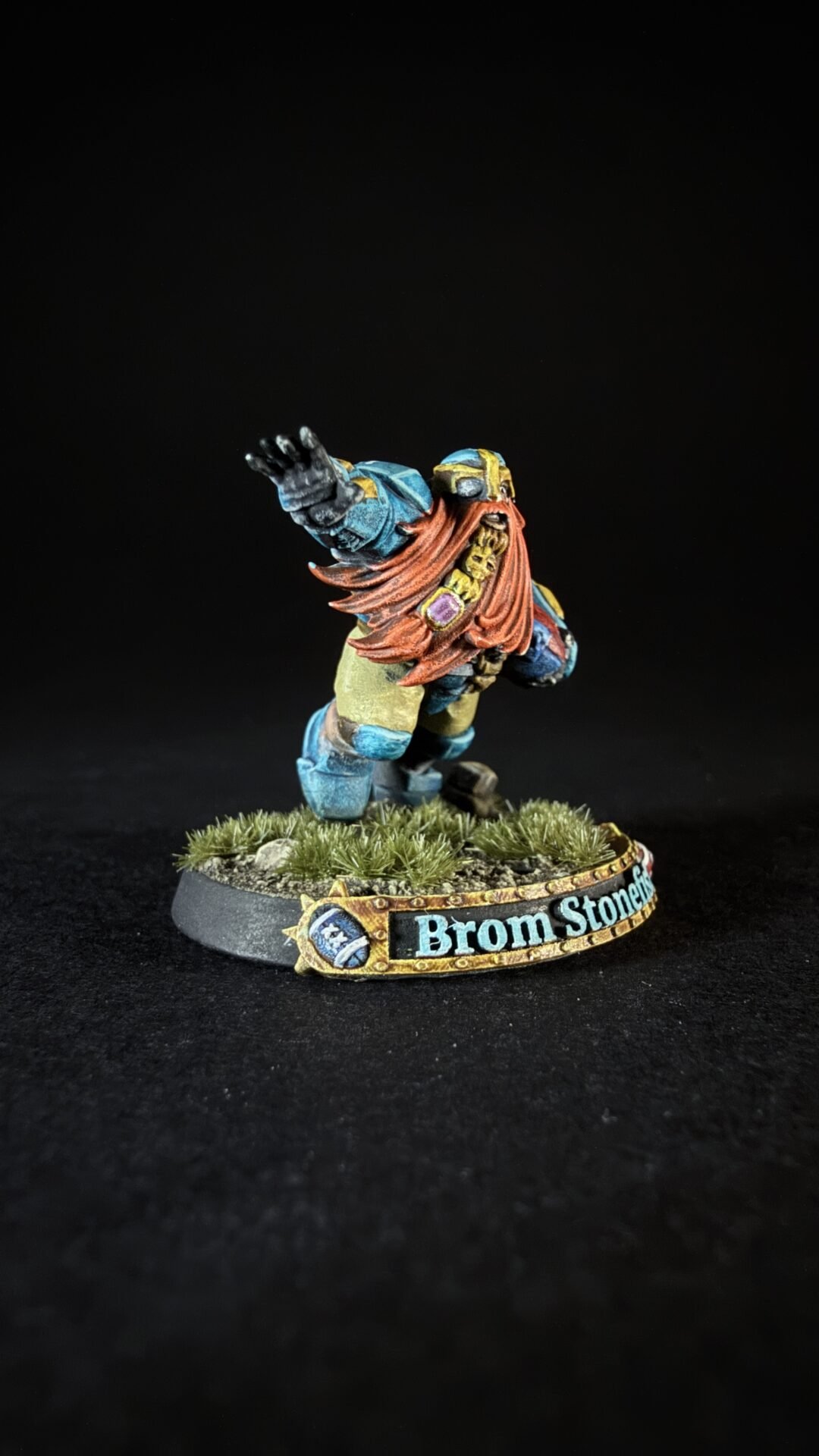 Miniature dwarf warrior with long red beard and blue armor raises his arm on a grassy base labeled Brom Stonehide.
