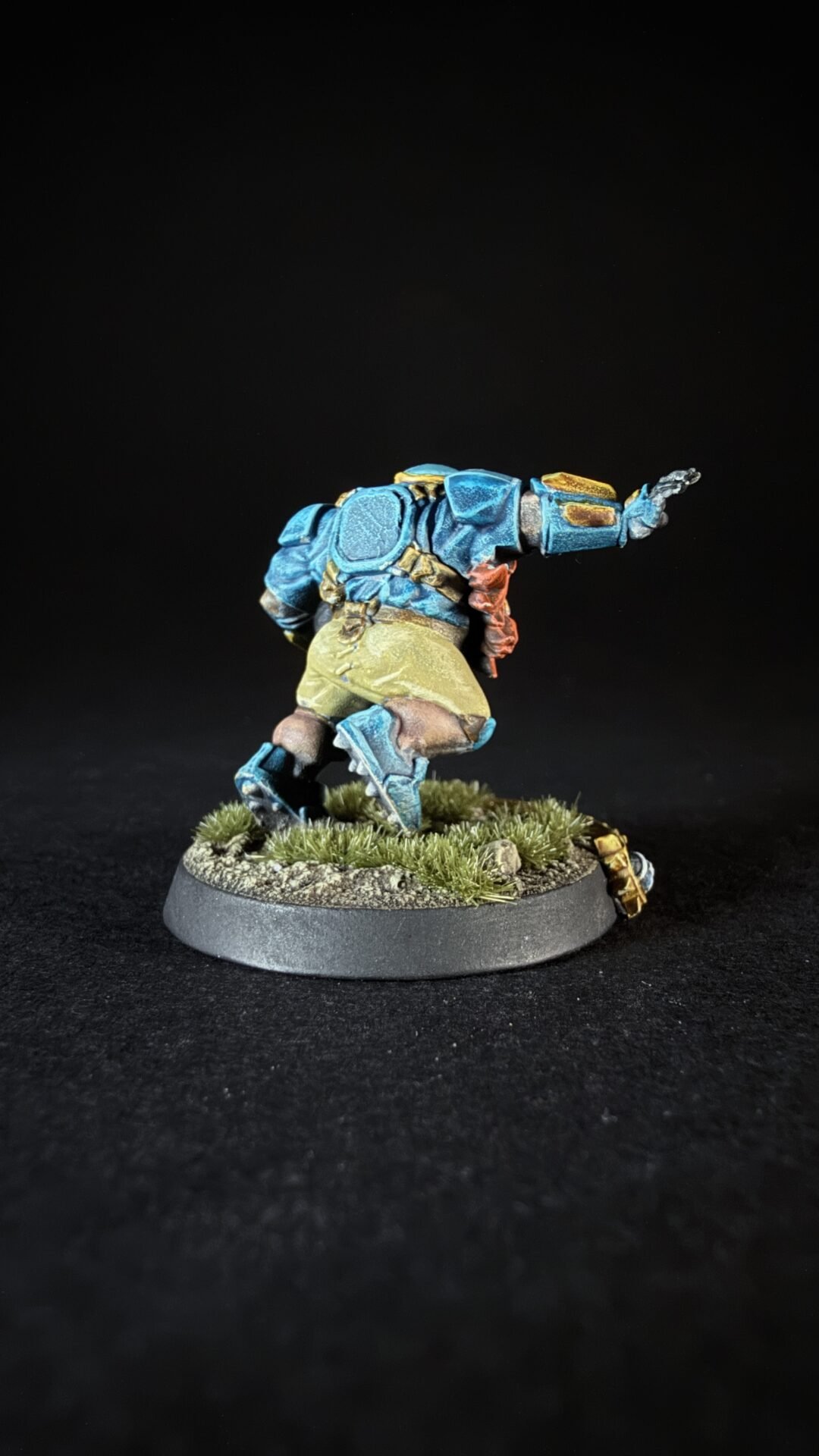 Painted sci‑fi miniature in blue armor leaping forward on a grassy base, with a brown belt and red sash detail against a black backdrop.