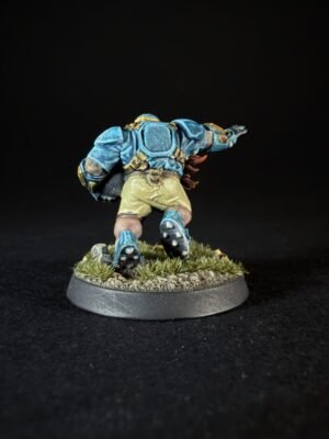 Back view of a fantasy miniature: a warrior in blue armor with gold accents, wearing beige shorts, standing on a grassy diorama base.
