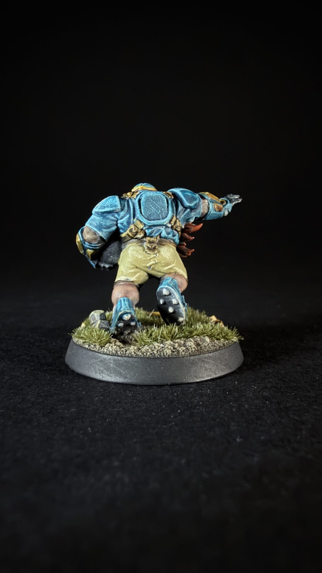 Back view of a fantasy miniature: a warrior in blue armor with gold accents, wearing beige shorts, standing on a grassy diorama base.