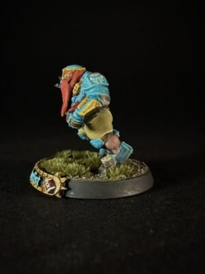 Colorful miniature figure in blue armor with a red scarf, built from blocky pieces, standing on a grassy round base against a black background.