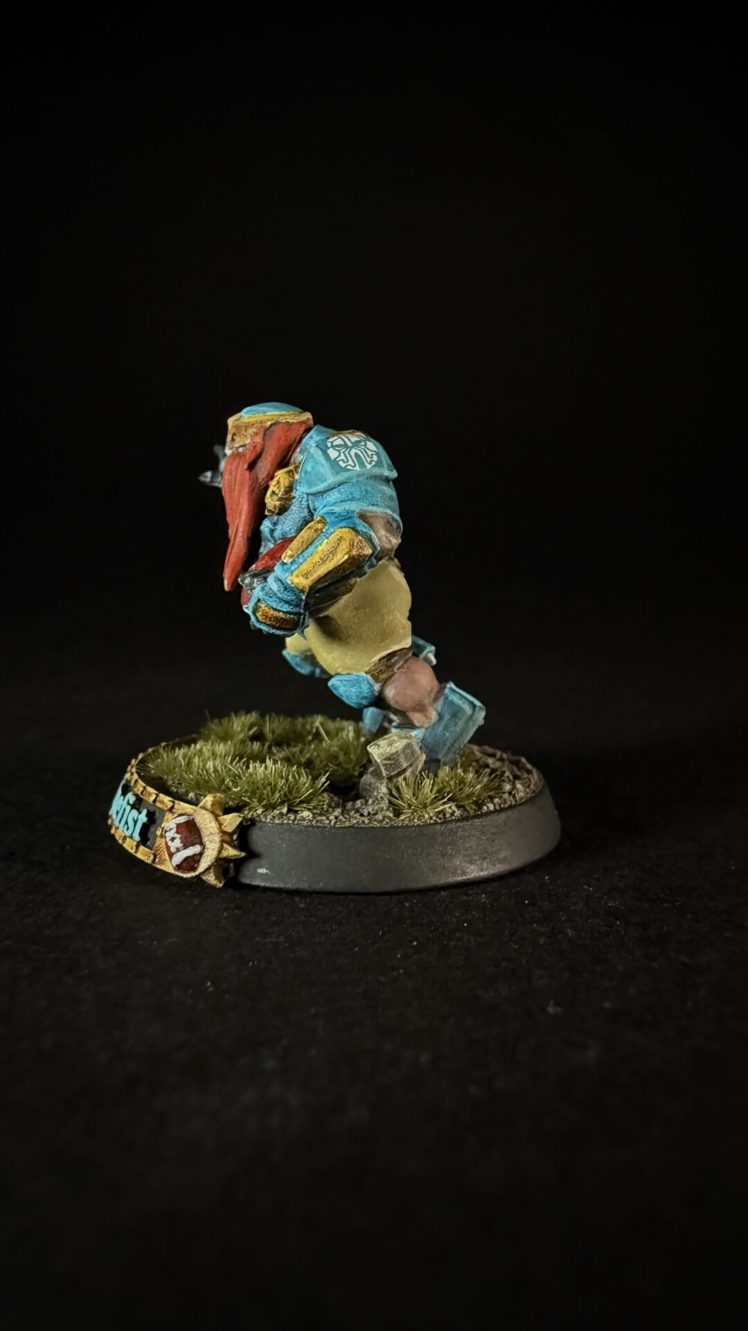 Colorful miniature figure in blue armor with a red scarf, built from blocky pieces, standing on a grassy round base against a black background.