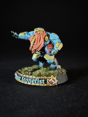 Colorful dwarf miniature in blue armor with a red beard, raising one arm, standing on a grassy base with a decorative banner reading 'From Stonefist'