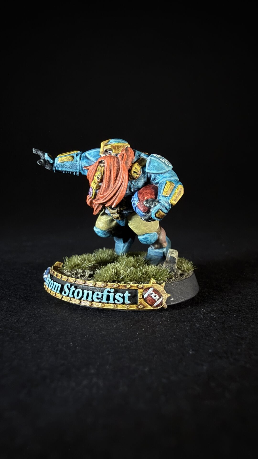 Colorful dwarf miniature in blue armor with a red beard, raising one arm, standing on a grassy base with a decorative banner reading 'From Stonefist'