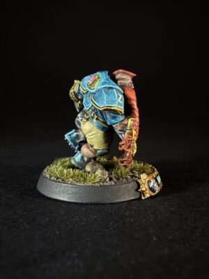 Tabletop miniature: blue armored warrior with yellow trim, standing on a grassy rocky base, red arm/weapon at the side.