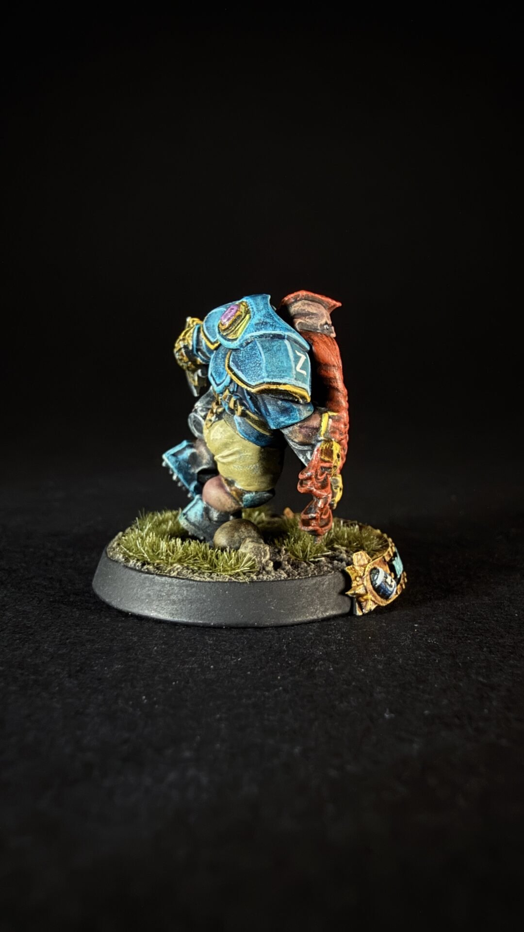 Tabletop miniature: blue armored warrior with yellow trim, standing on a grassy rocky base, red arm/weapon at the side.