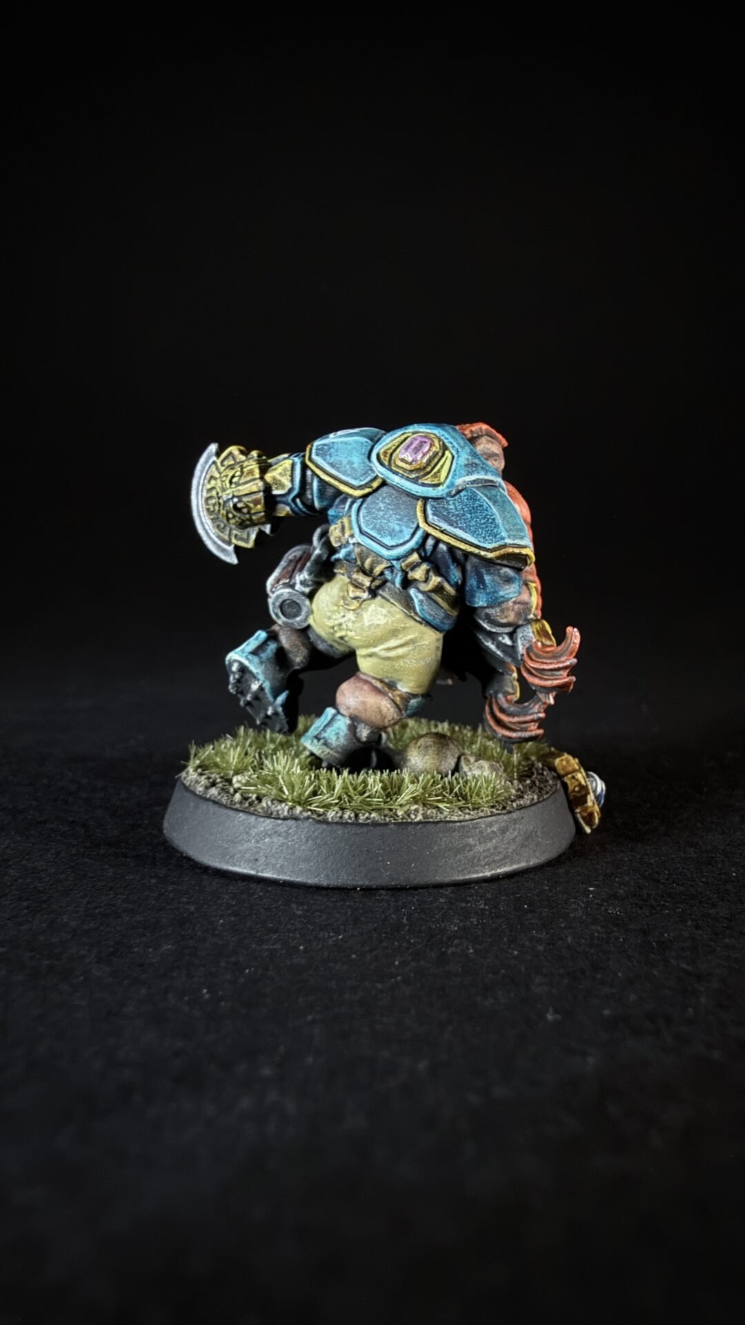 Back view of a painted fantasy miniature in blue armor with yellow trim, standing on a grassy base with a circular axe-like weapon nearby.