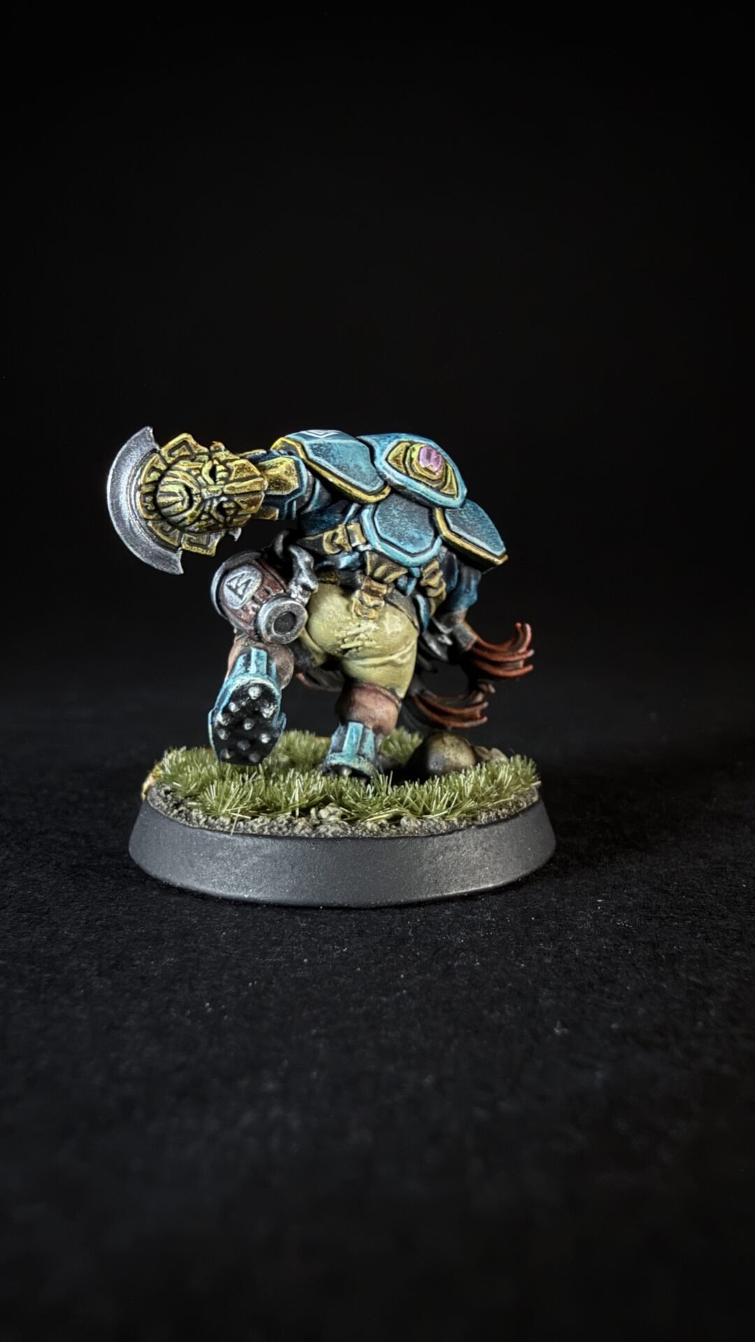 Painted fantasy miniature of a blue-armored orc-like warrior with a large weapon over its shoulder, standing on a grassy base.