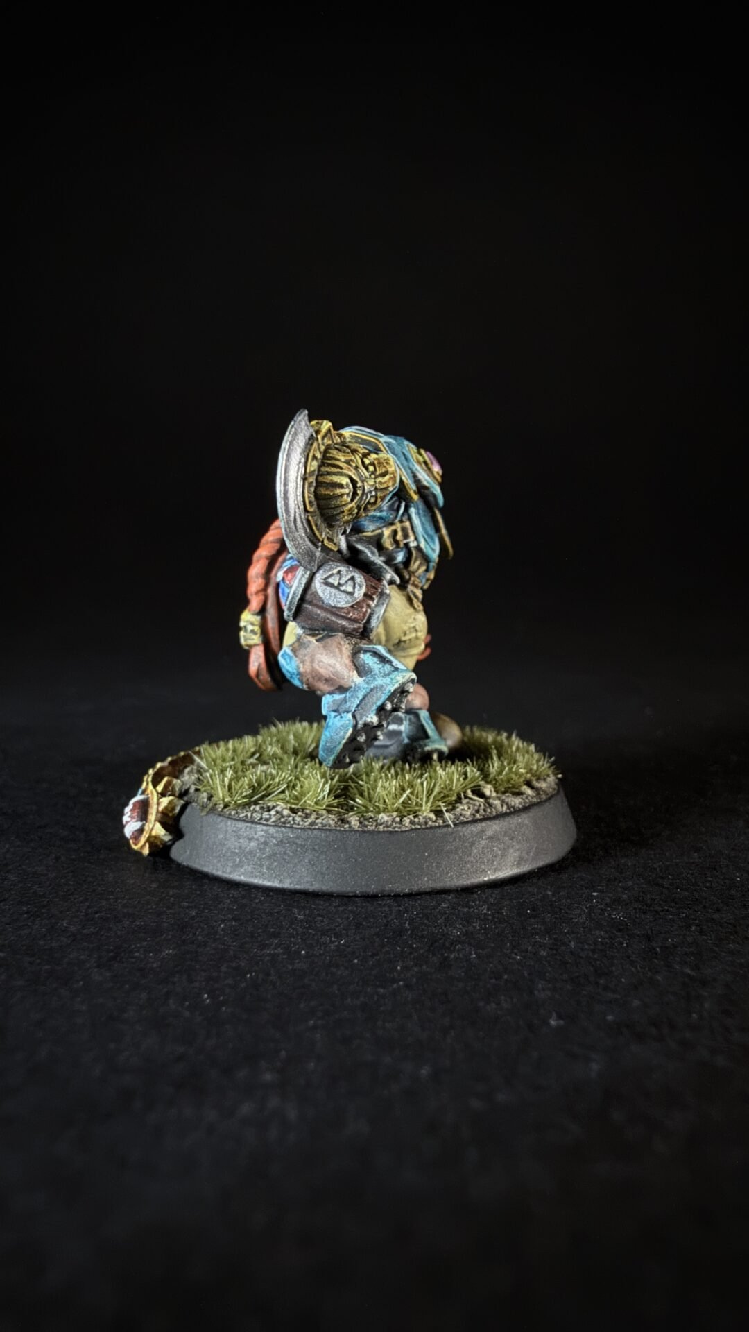 Miniature warrior with blue armor and red hair, holding a shield, standing on a grassy base against a dark background.