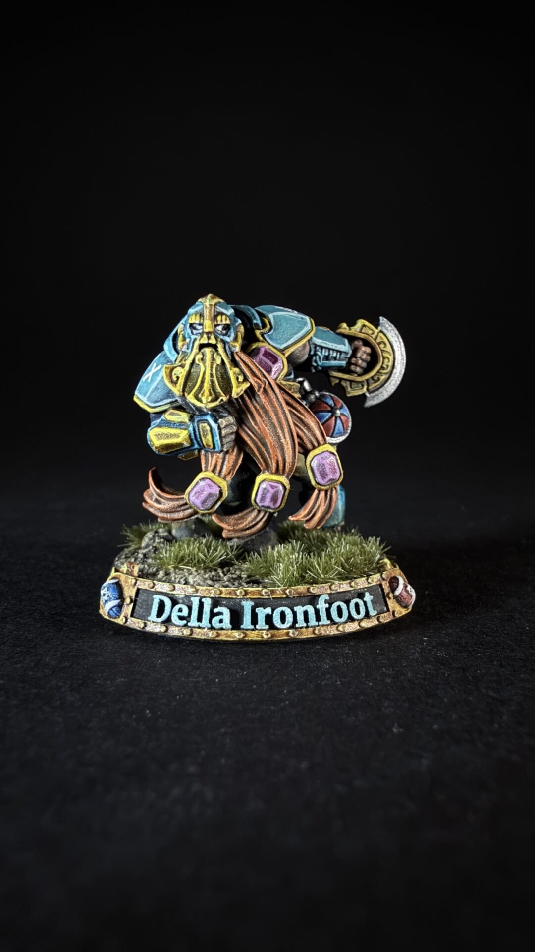 Painted fantasy dwarf miniature in blue armor with a long golden beard, wielding a hammer, on a grassy base labeled 'Della Ironfoot'.