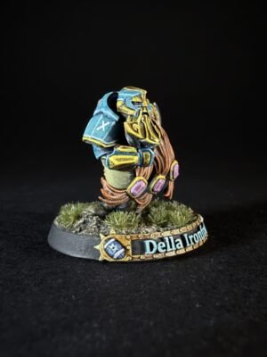 Painted fantasy miniature: a dwarf warrior in blue and gold armor with purple gems, standing on a rocky, grassy base. The plaque reads 'Della Ironf'.