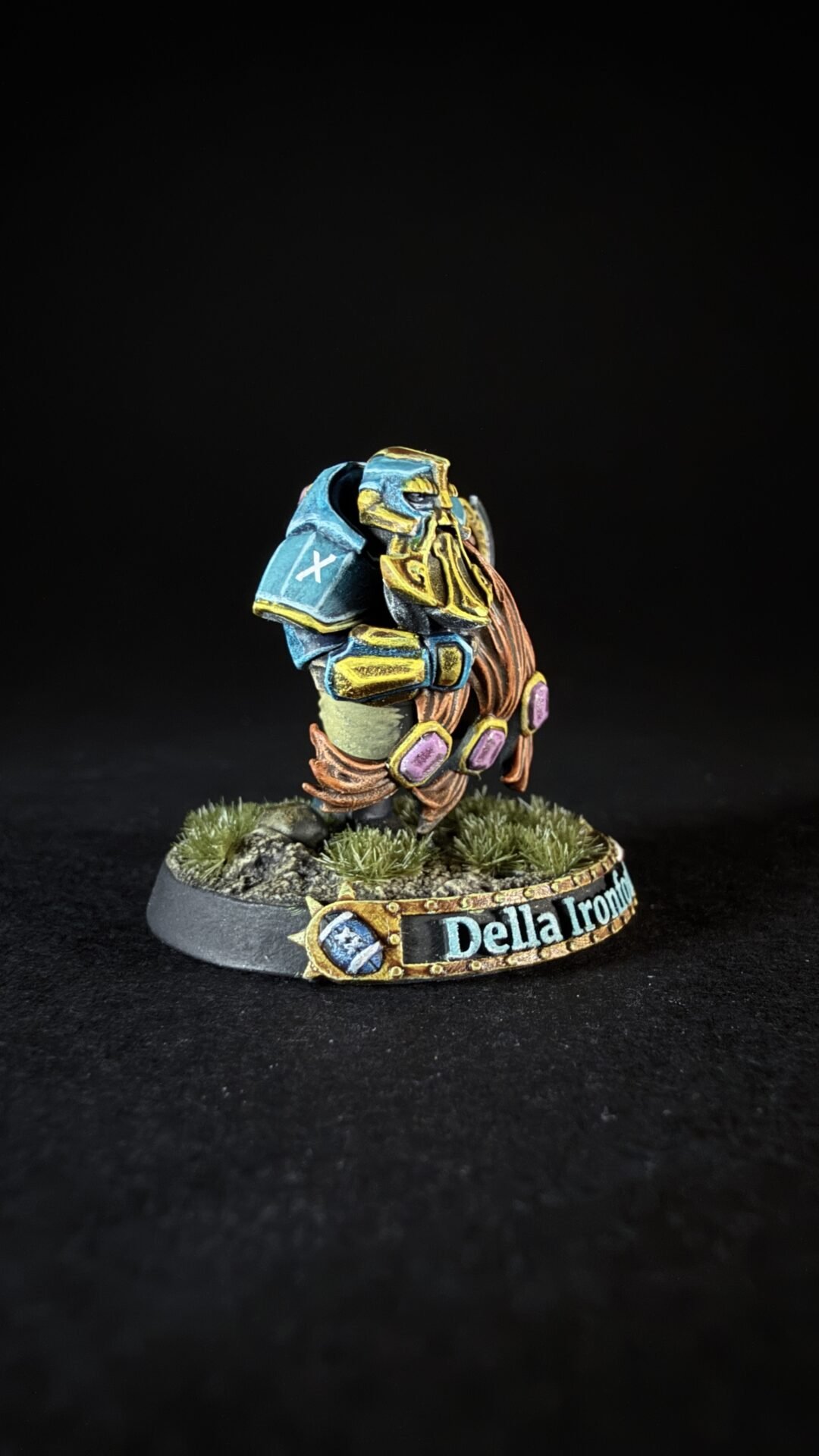Painted fantasy miniature: a dwarf warrior in blue and gold armor with purple gems, standing on a rocky, grassy base. The plaque reads 'Della Ironf'.
