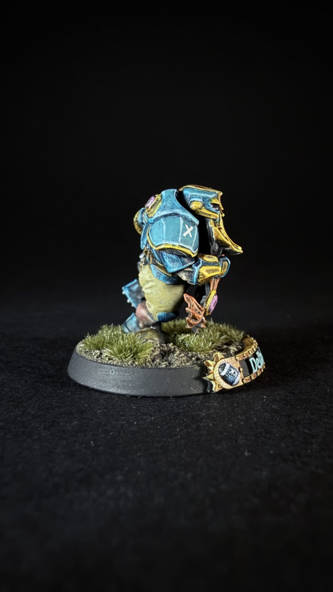 Small fantasy miniature figurine in blue armor with yellow trim, standing on a grassy diorama base against a black background.