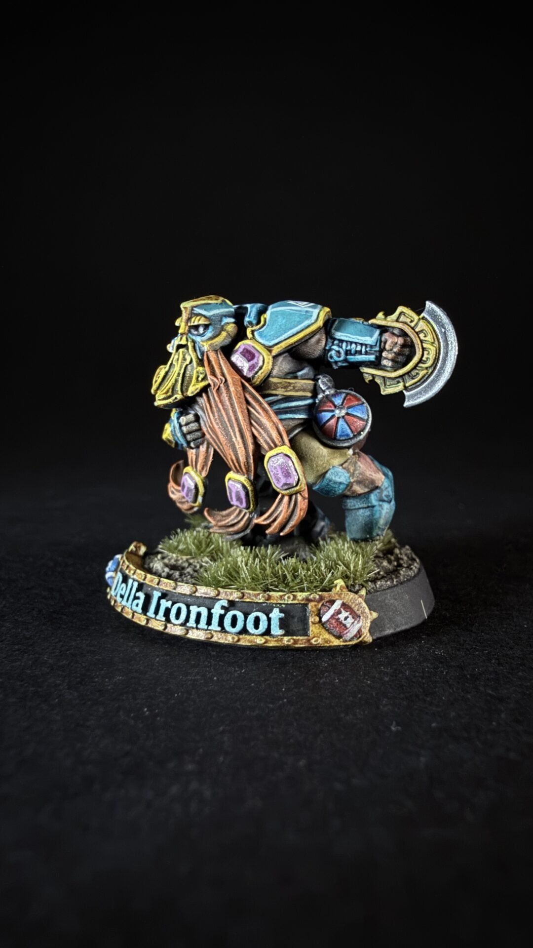 Della Ironfoot miniature: a colorful dwarf warrior in blue and gold armor, wielding a large ornate battle axe, standing on a grassy base with a banner bearing the name 'Della Ironfoot'.