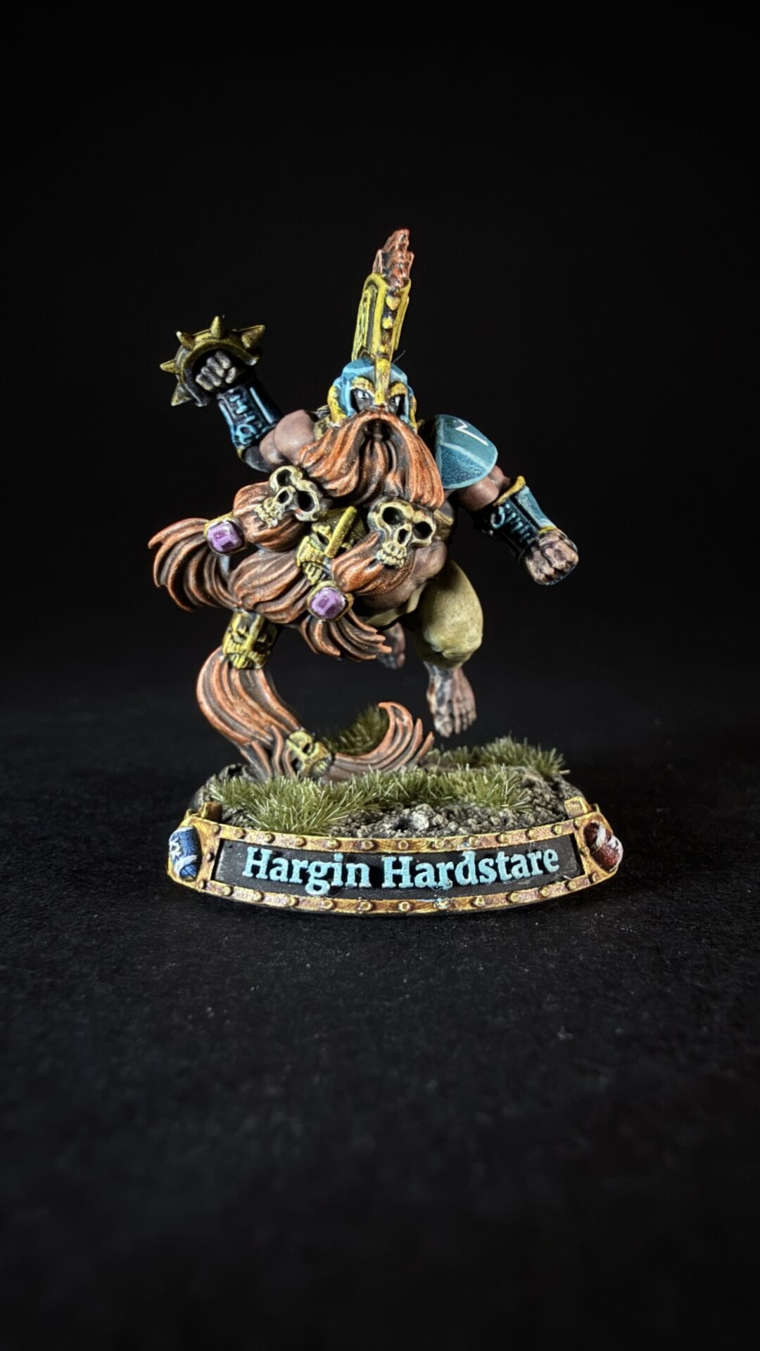 Painted fantasy miniature of a muscular warrior with skulls, braided orange hair, and blue armor, on a grassy base labeled 'Hargin Hardstare.