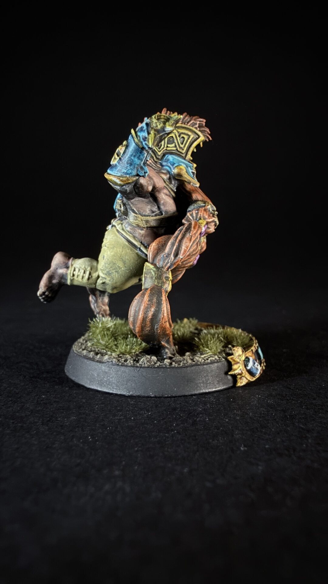 Colorful fantasy orc miniature charging forward, armed with a blue-and-gold shield, on a rocky grassy base.