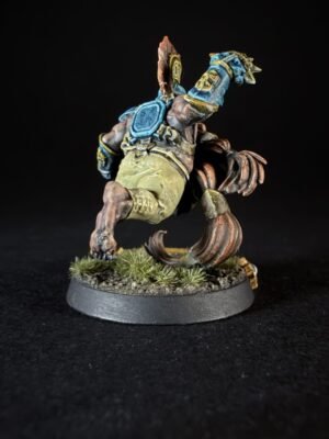 Brown, reptilian-like fantasy miniature in blue armor raises a spiked left arm, standing on a textured base with grass tufts.