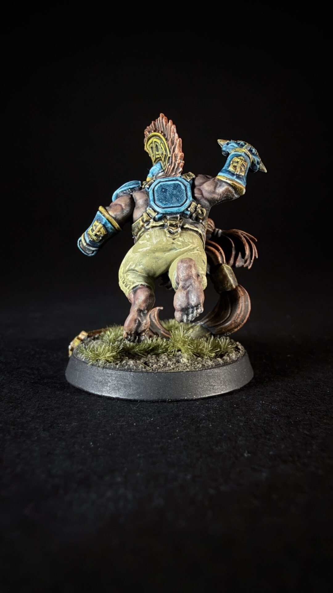 Back view of a painted fantasy miniature: muscular figure in blue armor with a feathered crest, tan shorts, and dynamic pose on a grassy base.