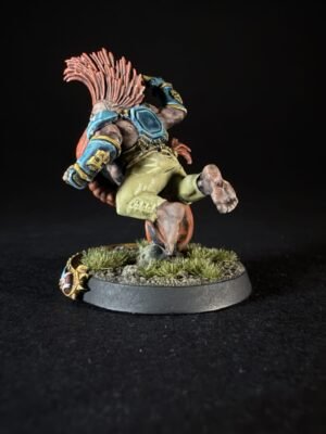 Fantasy miniature figure in blue armor and sandy shorts, mid-leap with red mane, on a textured grassy base.