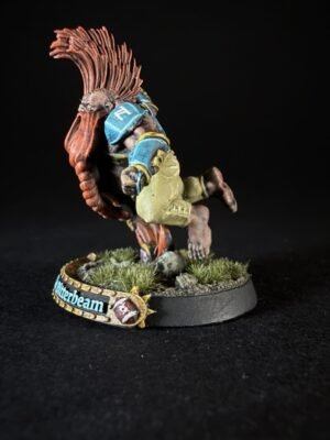 Miniature humanoid creature with a tall red crest, blue armor, and beige clothing, standing on a grassy base with a decorative banner nearby.