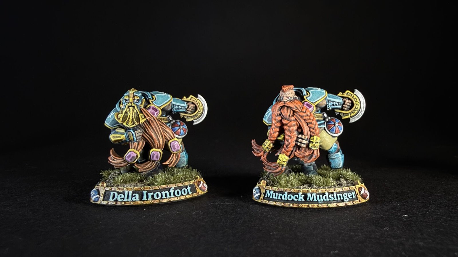 Two colorful fantasy dwarf miniatures on grassy bases, facing opposite directions, with name banners reading 'Della Ironfoot' and 'Murdock Mud­singer' (or similar).
