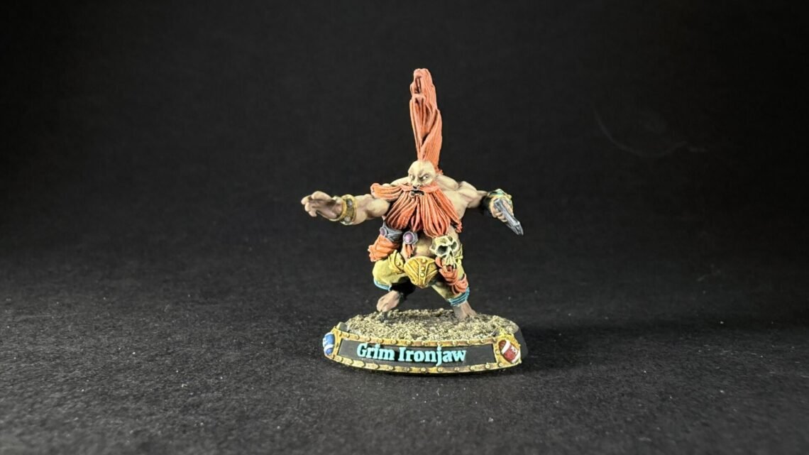 Miniature fantasy orc warrior Grim Ironjaw with tall orange hair plume, outstretched arms, detailed armor, standing on rocky base labeled Grimg Ironjaw (sic).