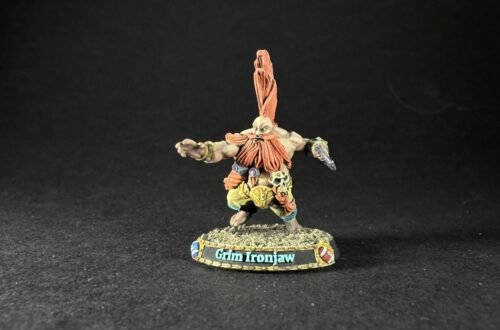 Miniature fantasy orc warrior Grim Ironjaw with tall orange hair plume, outstretched arms, detailed armor, standing on rocky base labeled Grimg Ironjaw (sic).