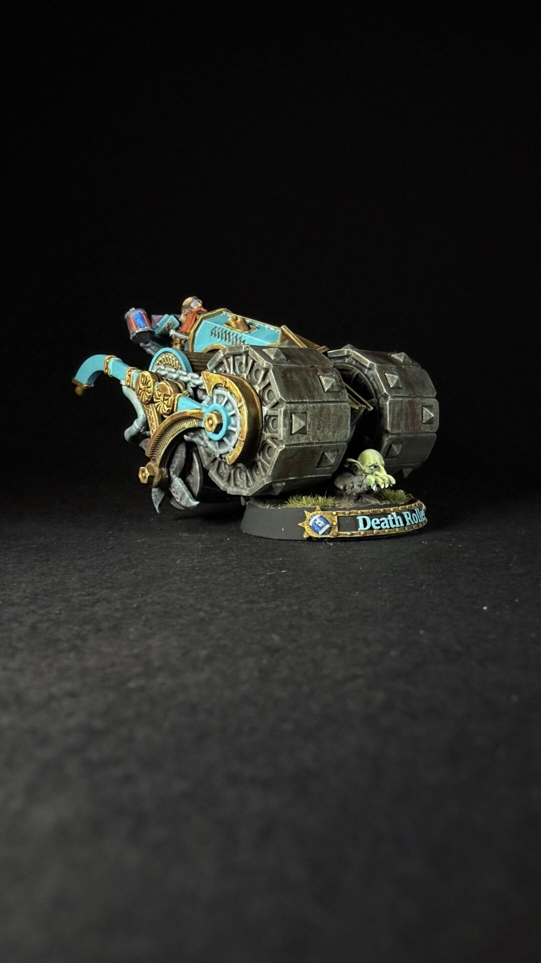Painted Warhammer model: turquoise and gold armored tank with large tread wheels, a small goblin-like pilot on top, set on a round base with 'Death Roll' lettering and tufts of grass.