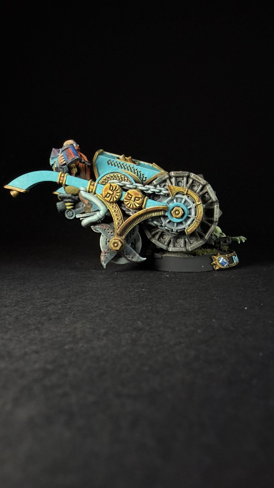 A turquoise and gold fantasy chariot miniature with a rider, ornate clockwork wheels, and mechanical blades on a black backdrop.