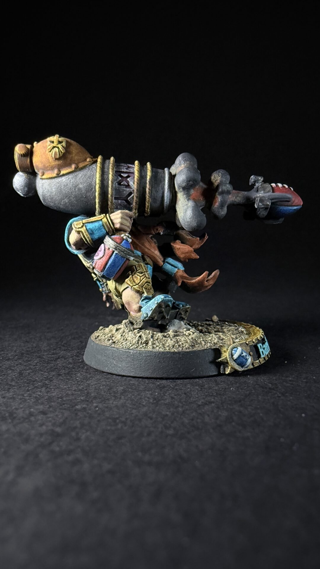 Painted dwarf artillery miniature firing a oversized cannon, colorful armor, standing on a rocky base against a black background.