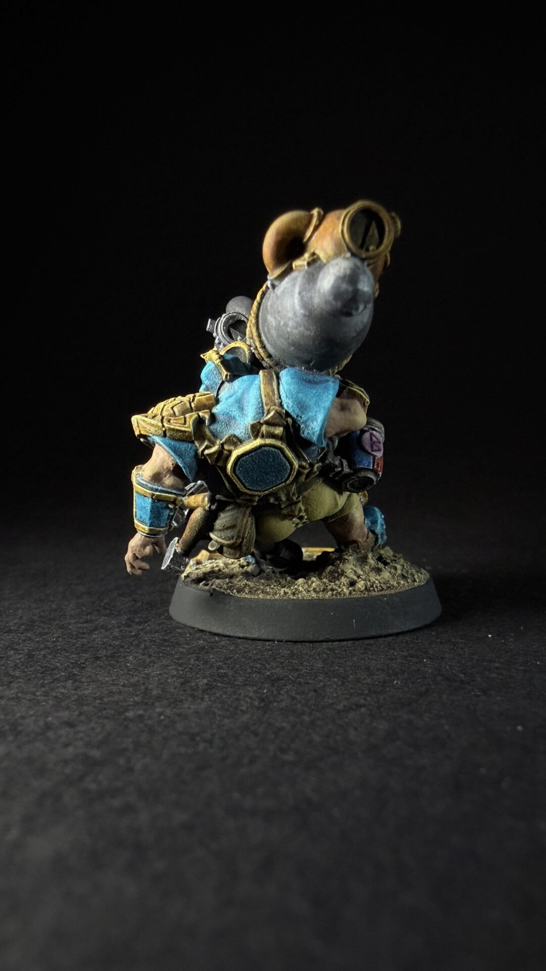 Back view of a small fantasy dwarf in blue and gold armor, standing on a rocky base against a black background. The figure is detailed with plates, straps, and a helmet-like headpiece, presenting a heroic, armored silhouette.