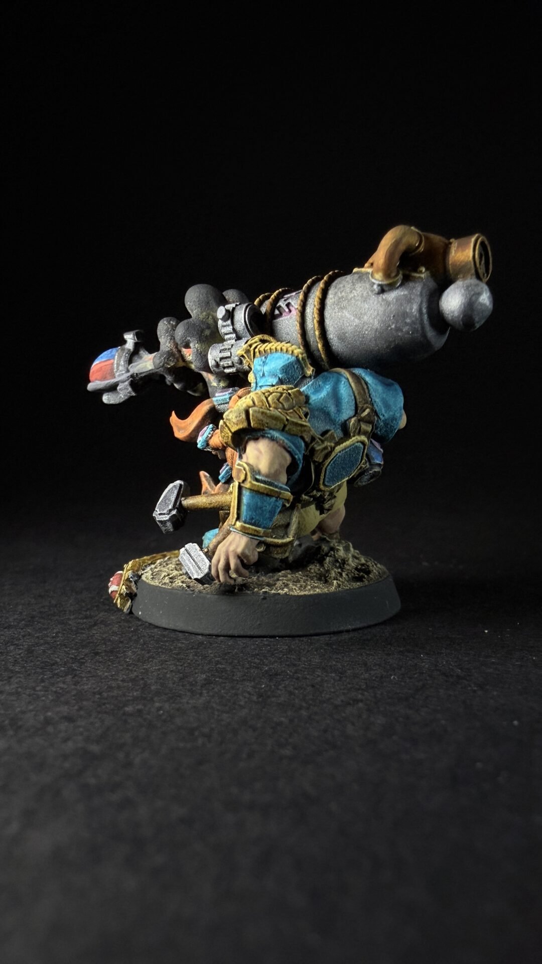 Dwarf warrior in blue and gold armor kneels on a rocky base, carrying a large steampunk-style cannon strapped to his back tied with ropes.