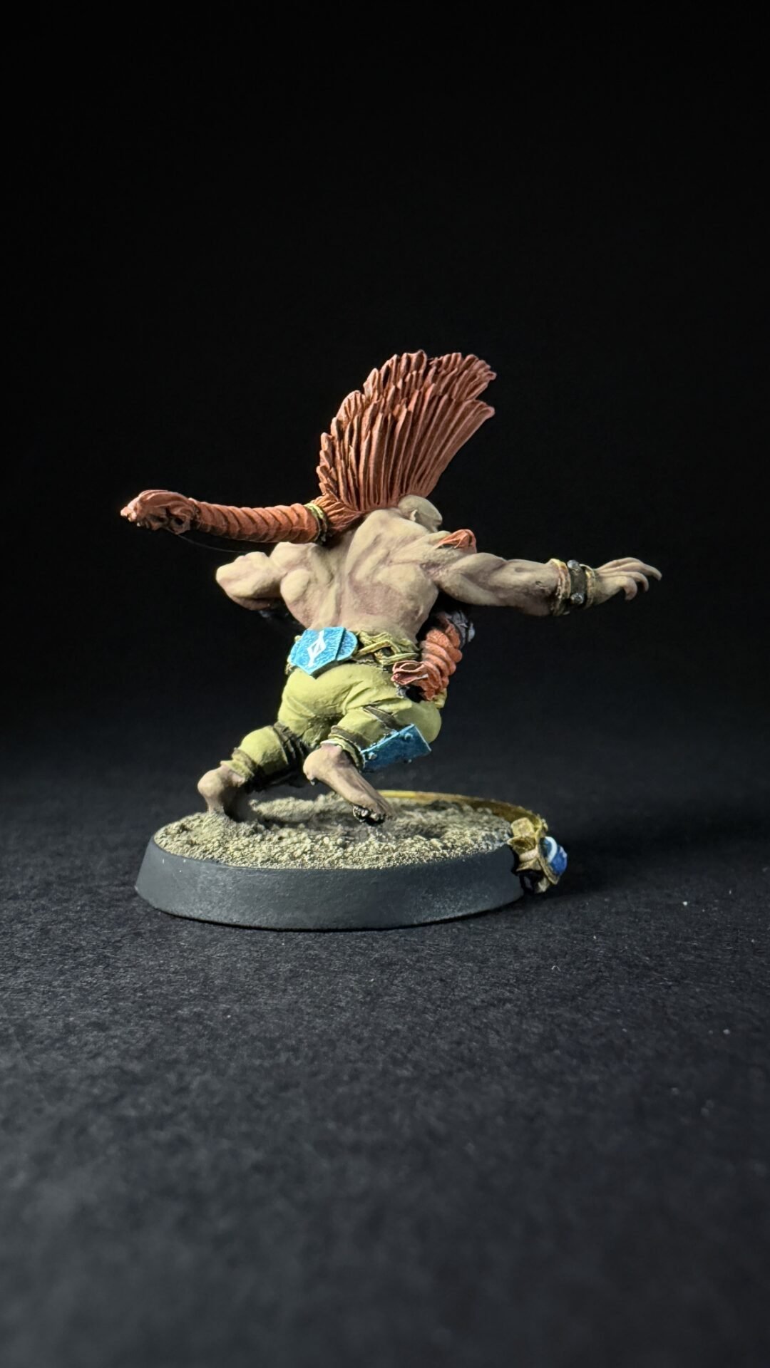 Back view of a running fantasy troll miniature, orange braided hair, gray-green skin, wearing green shorts and blue accessories on a textured base.