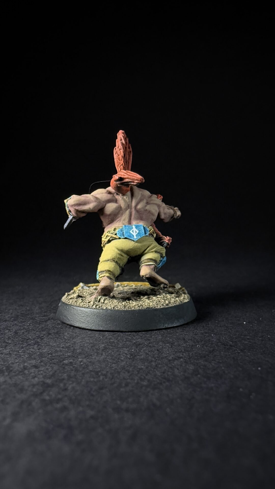 Back view of a miniature figure with orange hair tied in a plume, wearing olive pants and a blue belt on a rocky base against a dark background
