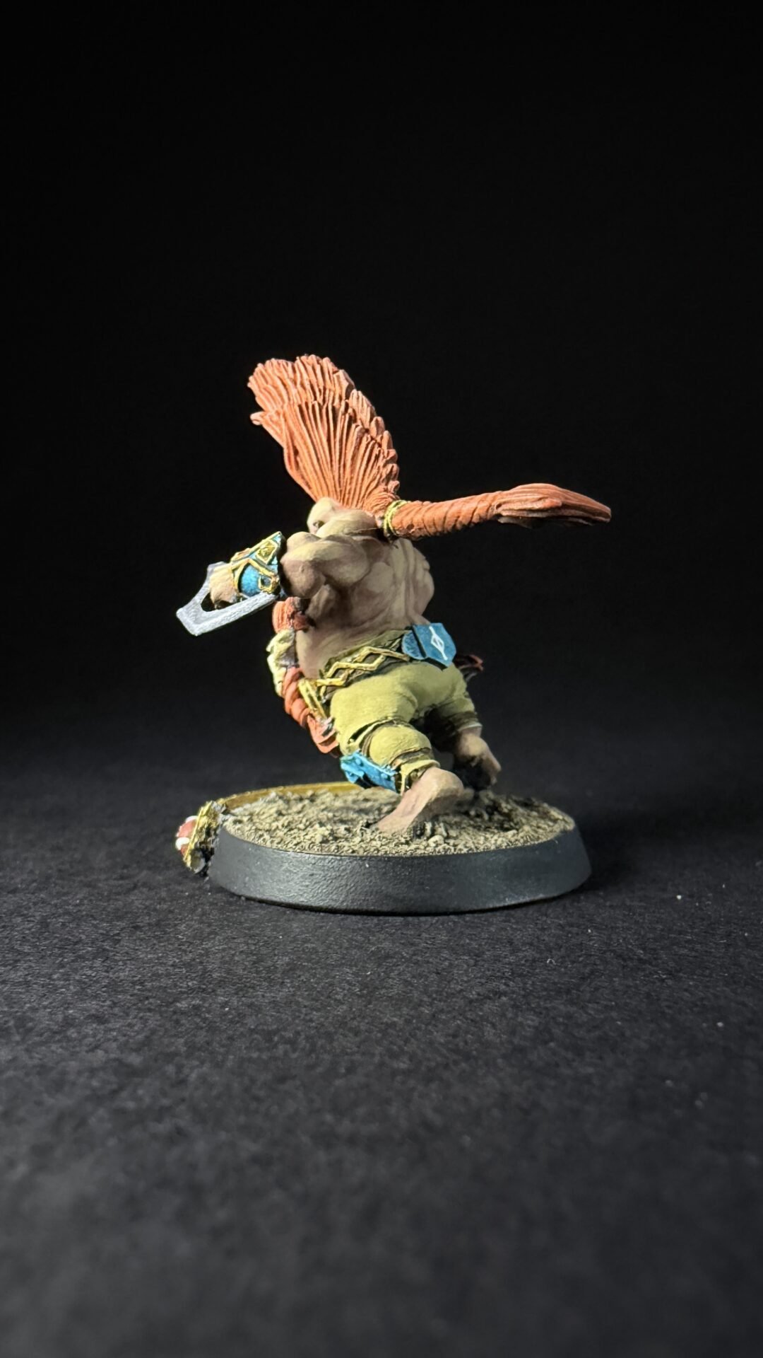 Miniature fantasy figure with orange feathered crest, muscular torso, green pants, and a curved blade, posed on a rocky base.