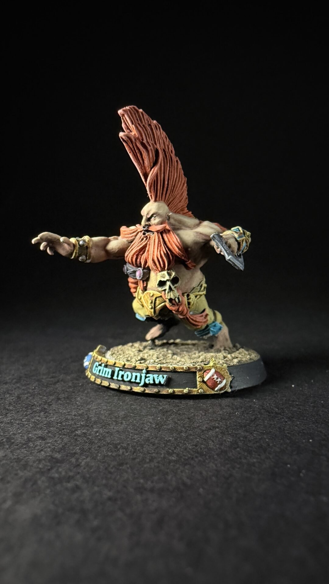 Fantasy orc warrior miniature with a tall orange mohawk, skull belt, and outstretched arms on a textured base reading 'Glin Ironjaw'.