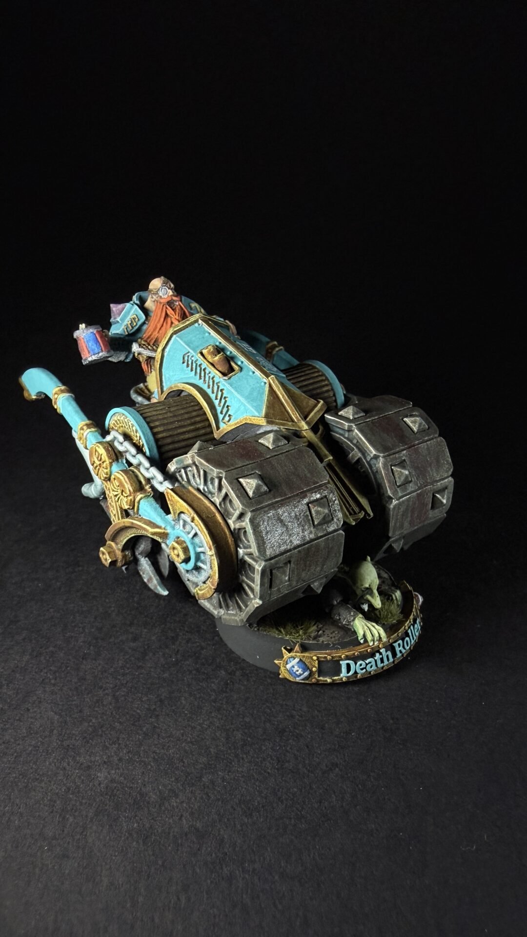 Warhammer Death Roller miniature with teal and gold armor, chained wheels, and a driver atop the siege‑engine on a black background.