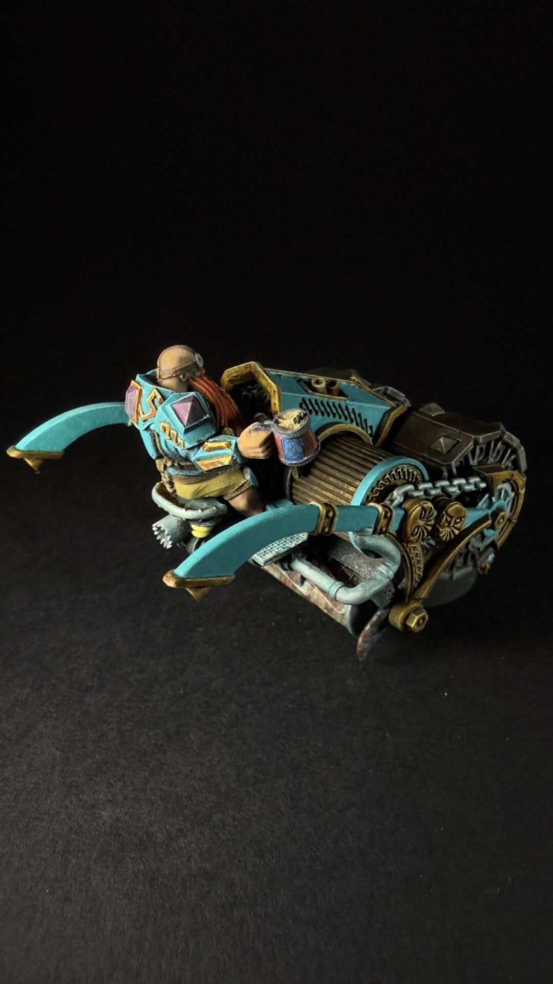 Teal and gold sci‑fi miniature vehicle with a driver perched on top, set against a black backdrop.