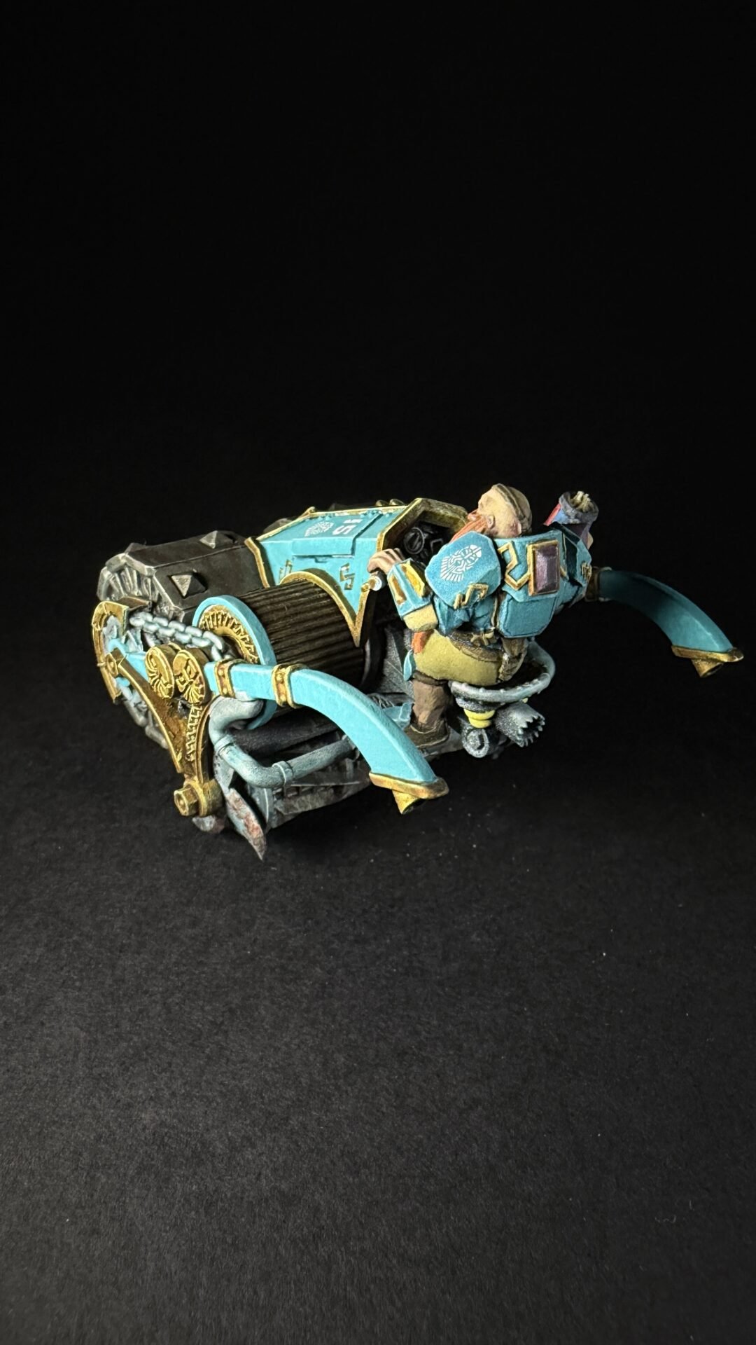 Teal and gold-painted sci‑fi miniature war machine with curved blades and ornate armor on a black background.