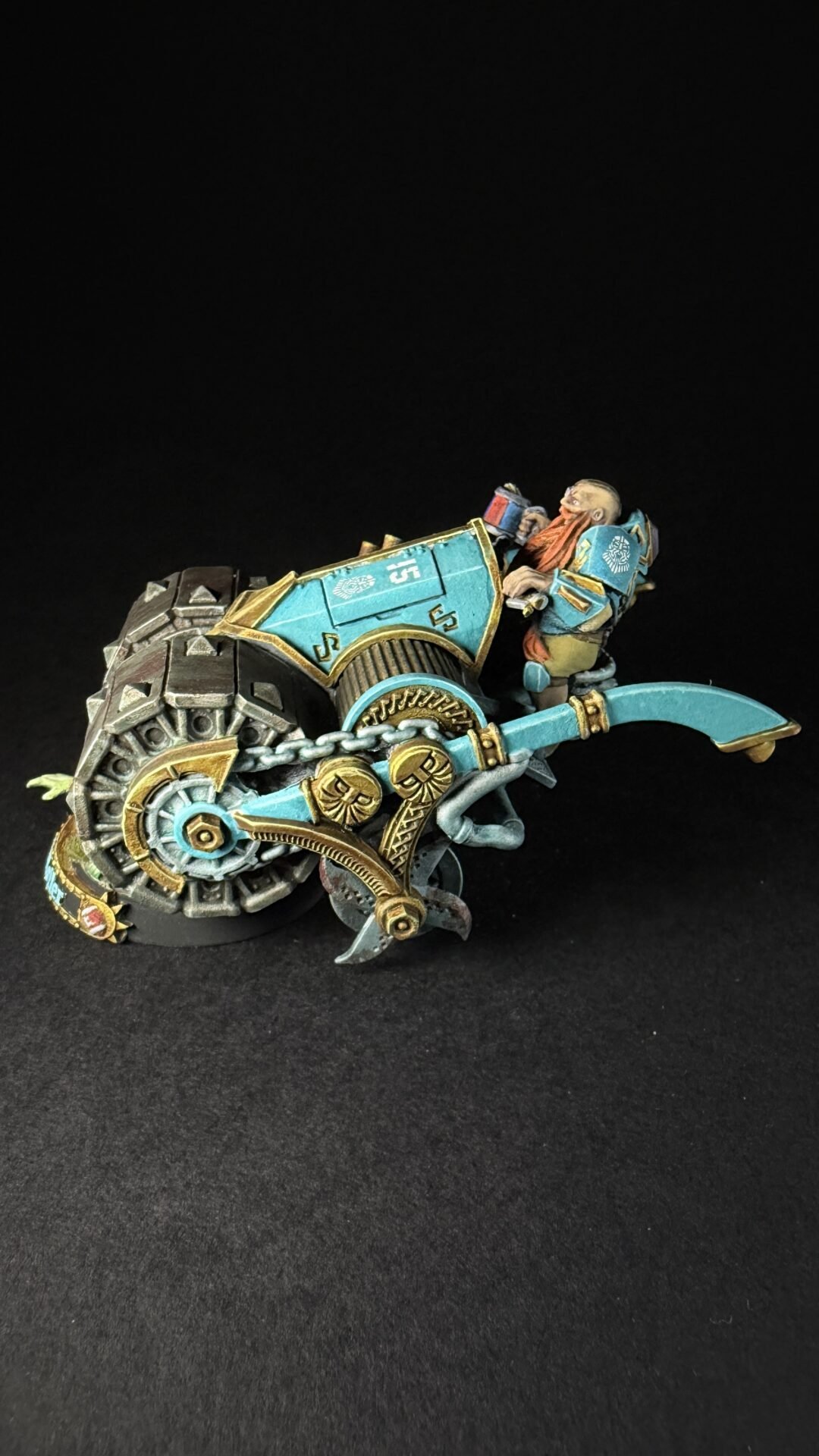 Miniature steampunk bike with teal and gold armor, ridden by a bearded dwarf against a black backdrop. Bearded dwarf pilot on a teal, gear-filled steampunk motorcycle miniature. Fantasy miniatures: a teal-and-gold riding vehicle with exposed gears and a bead-dwarf chauffeur. Teal and gold steampunk motorcycle model with a dwarf rider, detailed gears and chains.
