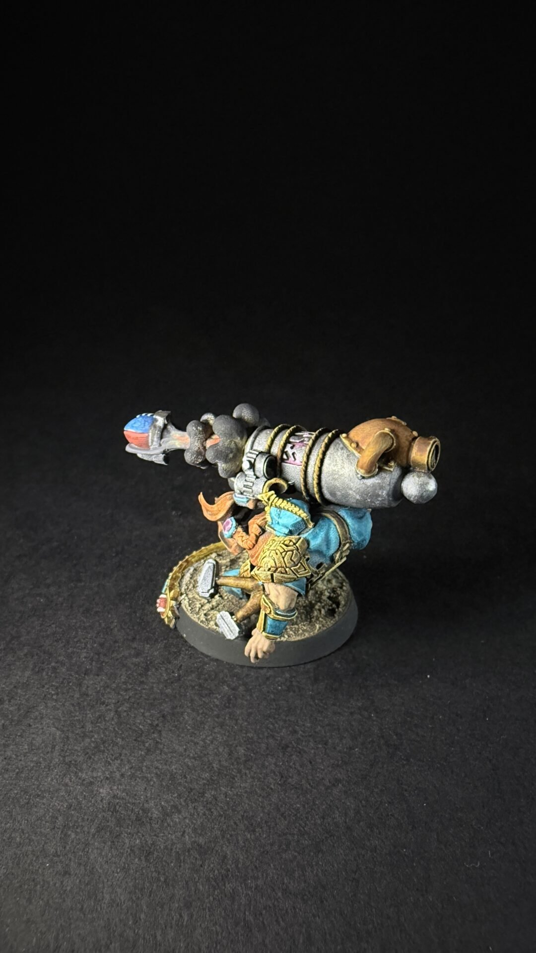 Painted fantasy miniature of a dwarf warrior with a large ornate cannon strapped to his back, wearing blue and gold armor and standing on a textured round base.