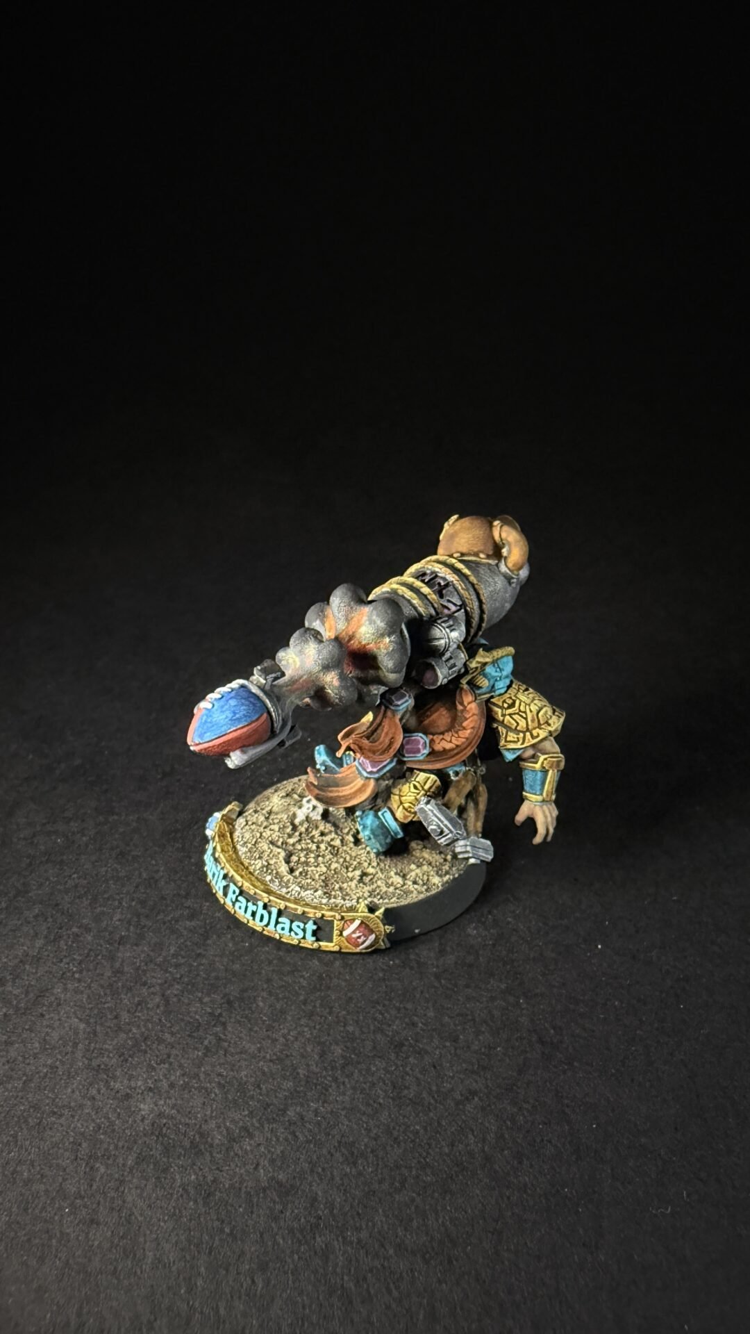 Painted tabletop miniature of a armored warrior with a teal and gold outfit, carrying a large lizard-like creature strapped to his back on a rocky base.
