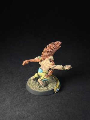 Fantasy miniatures figure: a muscular, bare-chested humanoid with an orange feathered headdress, lunging forward on a rocky base with decorative beads and armor pieces on wrists and waist.