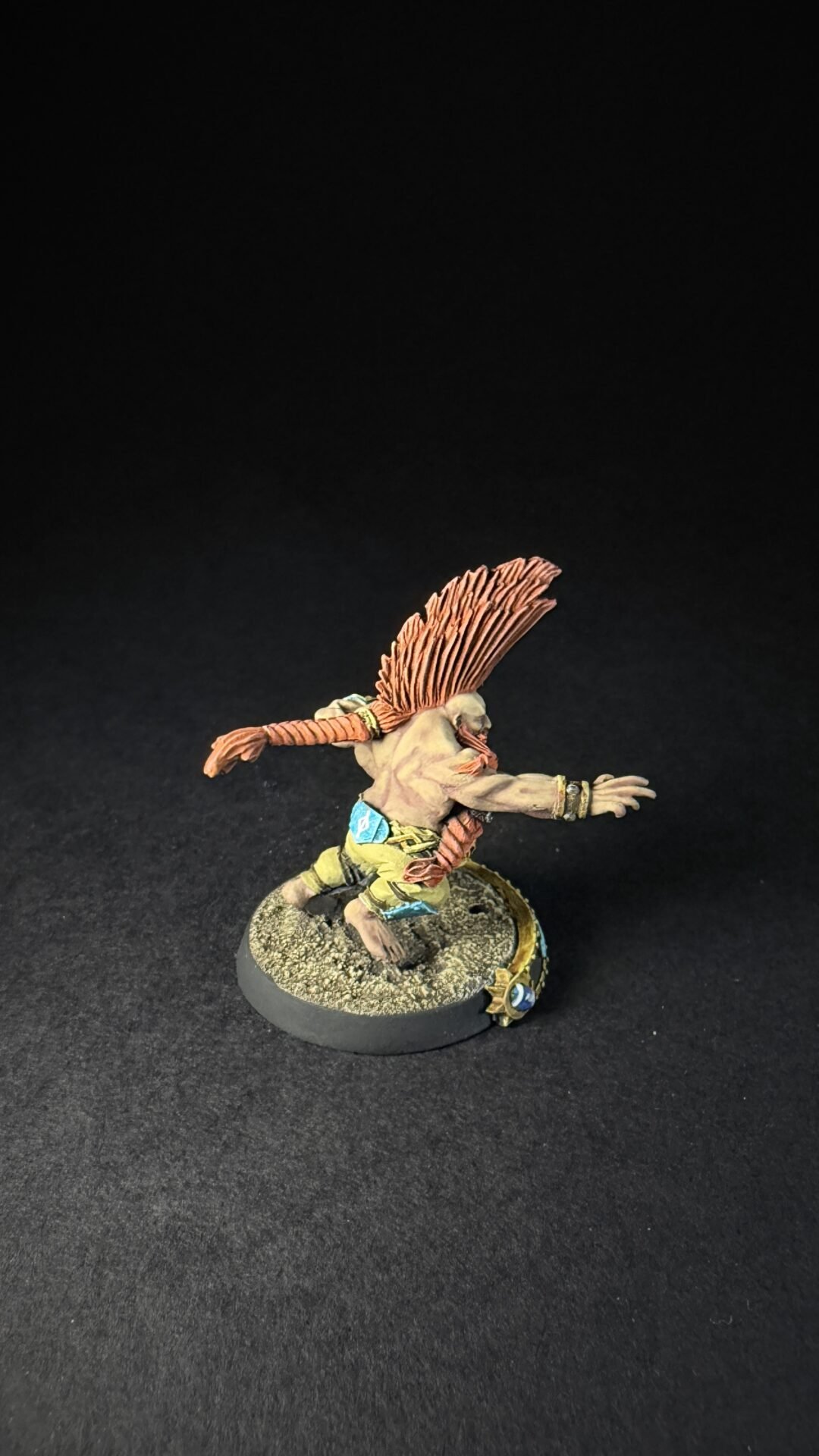 Fantasy miniatures figure: a muscular, bare-chested humanoid with an orange feathered headdress, lunging forward on a rocky base with decorative beads and armor pieces on wrists and waist.