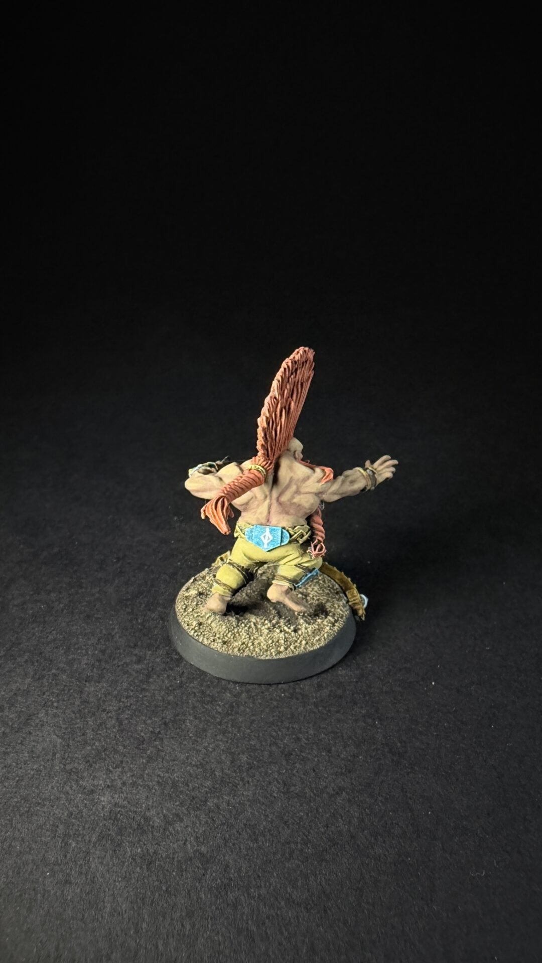 Front-facing miniature of a muscular fantasy orc with a tall orange feather headdress, outstretched arms, wearing a belt and tunic on a rocky base.